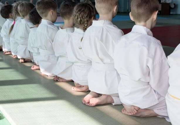 Children sit in a line wearing kimono and white belts for beginner level