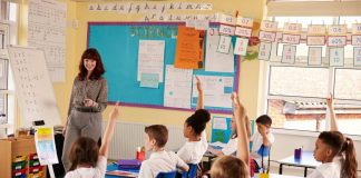 Teacher leading a lesson in a UK primary classroom while pupils in uniform sit at desks with their hands raised
