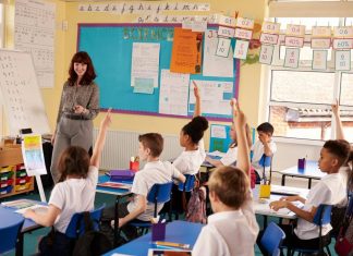 Teacher leading a lesson in a UK primary classroom while pupils in uniform sit at desks with their hands raised