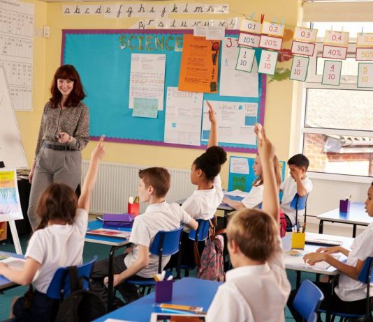 Teacher leading a lesson in a UK primary classroom while pupils in uniform sit at desks with their hands raised
